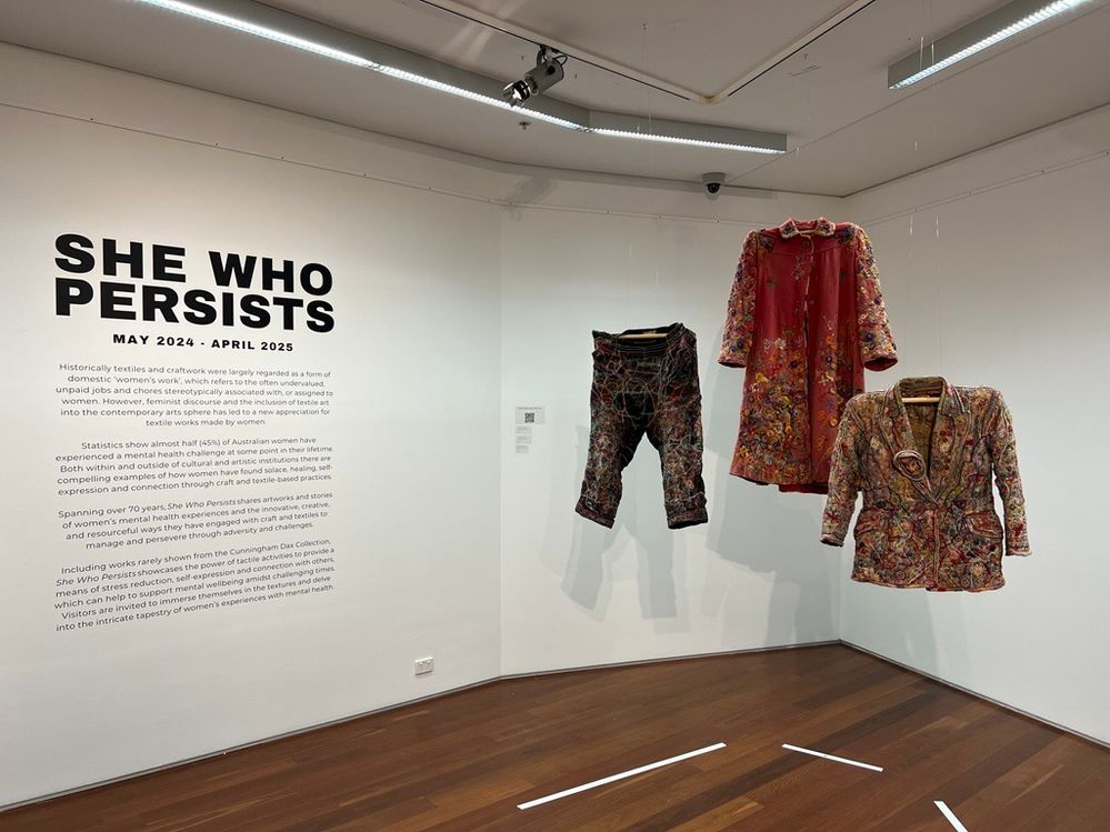 2)	She Who Persists installation image, 2024. Featuring artwork: Edith Agnes Harrington, Pink Twill Coat, c.1950-1960, woolen embroidered 		garment, 120 x 62cm, 2000.011 Edith Agnes Harrington, Short Jacket, c.1950-1960, grey serge embroidered 		garment, 70 x 60cm, 2000.012 Edith Agnes Harrington, Black Twill Trousers, c.1950-1960, black wool embroidered 	garment, 86 x 50cm, 2000.013. Courtesy of The Dax Centre.